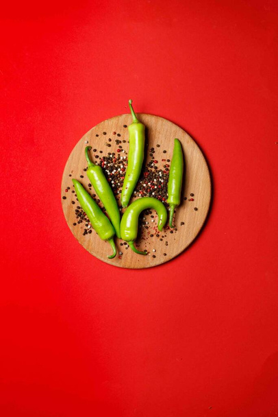 Zielone strąki chili na okrągłej drewnianej desce z kolorowym groszkiem pieprzu. Nad głową, na czerwonym tle, w środku kompozycji, pionowa rama, z przestrzenią - Zdjęcie, obraz