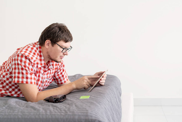 Koncepcja zakupów online. Młody mężczyzna leży na łóżku i robi zakupy online za pomocą tabletu - Zdjęcie, obraz