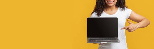 Offerta online. Irriconoscibile signora afro-americana che punta al computer portatile con schermo nero, sfondo giallo, spazio libero - Foto, immagini