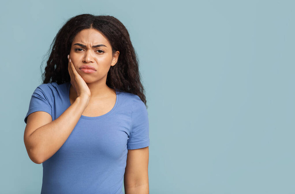 Schmerzhafte Zähne. Unzufriedene schwarze Dame mit Zahnschmerzen berührt Wange über blauem Hintergrund mit Freiraum - Foto, Bild