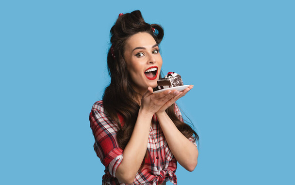 Charming pinup lady in retro wear eating chocolate cake on blue studio background - Photo, Image