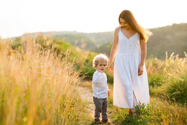 Hermosa mujer en un vestido camina en la naturaleza con su hijo con el pelo rizado - Foto, imagen