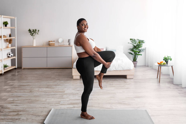Femme noire en surpoids étirant la jambe, faisant des exercices de flexibilité sur le tapis de yoga, menant un mode de vie actif pendant la cohabitation - Photo, image