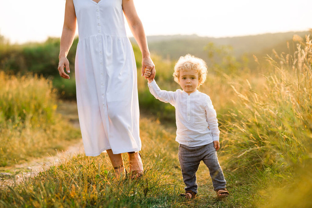 Όμορφη γυναίκα σε ανοιχτό λευκό φόρεμα οδηγεί το γιο της από το χέρι κατά μήκος του μονοπατιού ενός γραφικού τομέα - Φωτογραφία, εικόνα