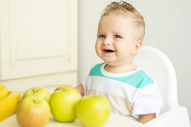 cute baby boy sitting at the table in child chair eating apple on white kitchen. - Foto, Imagen