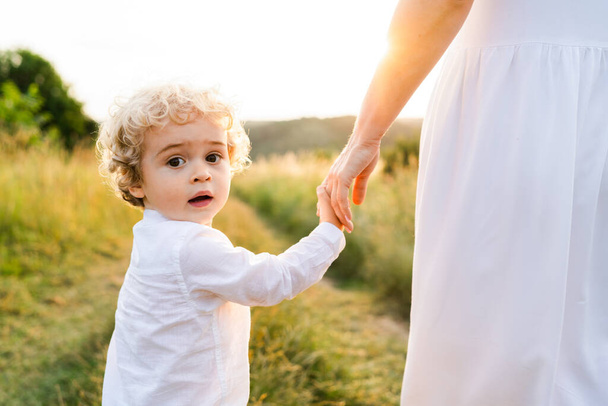 Cute boy with blond curly hair walks with his mother along a picturesque field at sunset and turns back looking at the camera - Zdjęcie, obraz