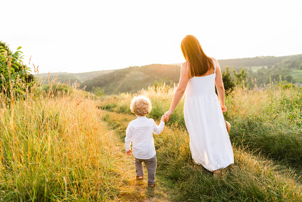 Anne ve oğlu gün batımında güzel bir yamaçta el ele yürüyorlar. - Fotoğraf, Görsel