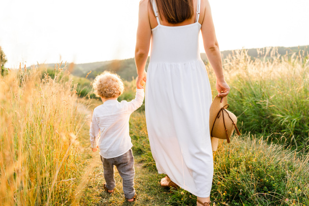 Mujer en un hermoso vestido blanco y su lindo hijo de pelo rizado están caminando en un campo de verano vista trasera - Foto, Imagen
