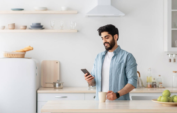 Hombre árabe joven feliz usando teléfono inteligente y beber café de la mañana en la cocina, espacio vacío - Foto, Imagen