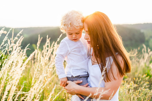 Loving mother holding her son in her arms while standing in a picturesque field at sunset - Fotografie, Obrázek