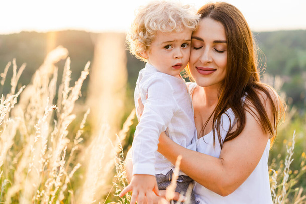 Loving mother hugs her son while standing in the middle of a picturesque field at sunset - Zdjęcie, obraz