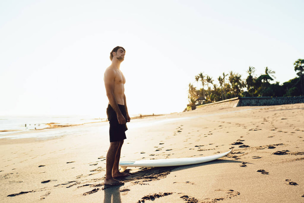 Side view full length sportive male surfer standing barefoot on sandy seashore near surfboard and stretching neck before going to surf - 写真・画像