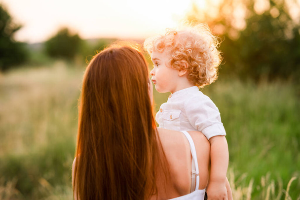 Menino bonito com cabelo encaracolado loiro nos braços de sua mãe enquanto caminha na natureza - Foto, Imagem
