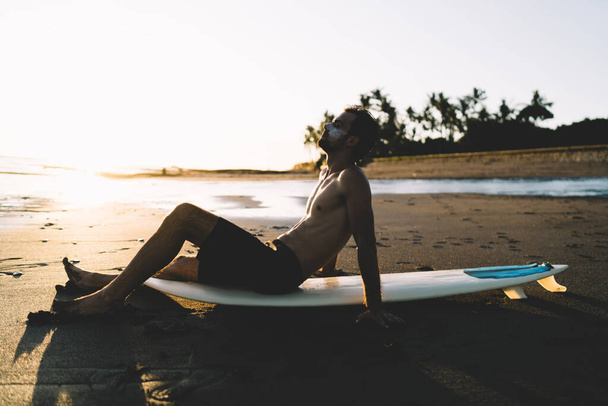 Side view of male person with professional surfboard looking for inspiration while sitting at sand beach during twilight time, young man surfer enjoying recreation near ocean during summer getaway - Foto, Bild