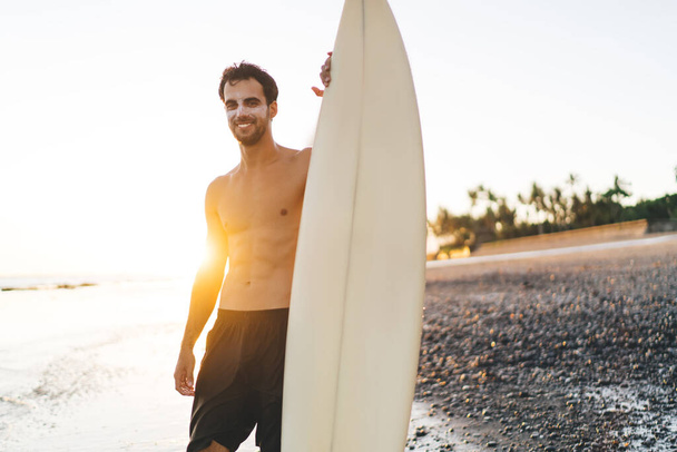 Half length of cheerful man with professional surfboard smiling at camera during weekend time for hobby practice, happy male surfer enjoying summer vacations at Malibu Beach in sunny California - 写真・画像