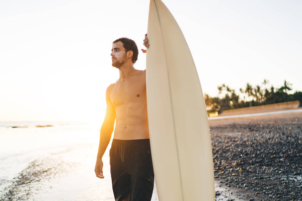 Young shirtless sportsman with sunscreen on face standing with surfboard on shore and looking away at sea on sunny evening - Foto, afbeelding