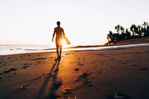 Rear view of sportive man walking at summer seashore with sunset spending day for practice hobby with surfboard, active male surfer enjoying travel vacations for catch wave in Dominicana Republic - Fotoğraf, Görsel