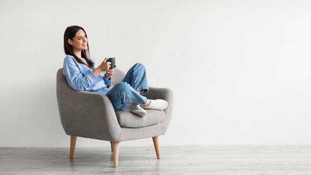 Fröhliche junge Frau trinkt aromatischen Kaffee, während sie im gemütlichen Sessel vor weißer Wand chillt, Panorama - Foto, Bild