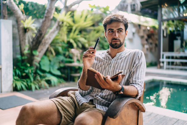 Pensive male writer in shirt and shorts sitting on cozy chair with open journal and raised pen on poolside near trees and looking away - Foto, Imagem