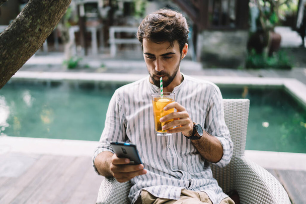 Bearded male in casual wear enjoying bright refreshing drink with lemon slice and straw while text messaging on mobile phone sitting on chair near swimming pool in cafeteria - Foto, Bild