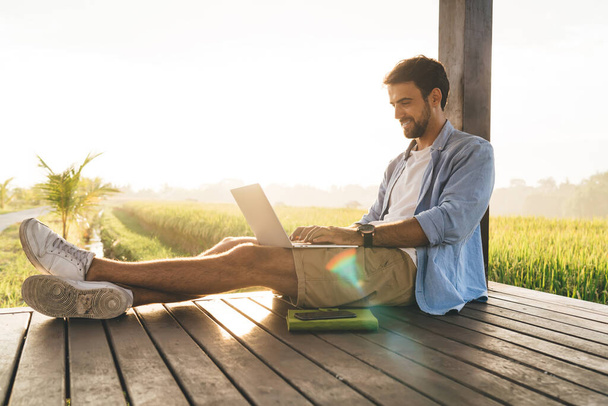 Side view of content male remote worker in casual wear browsing internet on netbook while sitting with crossed legs on wooden terrace near grass field in bright sunlight - Foto, imagen