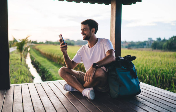 Jeune homme barbu en vêtements d'été décontractés surfant sur les médias sociaux sur téléphone portable et souriant joyeusement assis dans une pergola en bois près d'un champ vert dans un pays tropical - Photo, image
