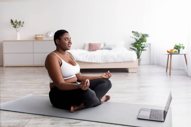 Tombul siyah bir kadın internette meditasyon ya da yoga kursuna dizüstü bilgisayar başında oturuyor, gözleri kapalı evde poz veriyor. - Fotoğraf, Görsel
