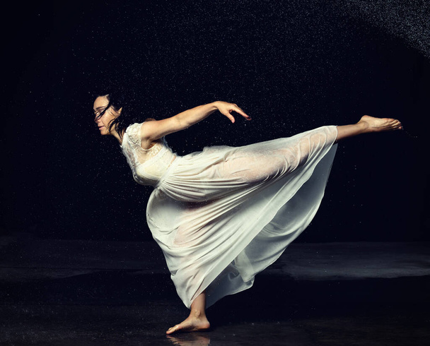 вродлива жінка з кавказького вигляду з чорним волоссям танцює у краплинах води на чорному тлі. На жінці біле шифонове плаття. - Фото, зображення