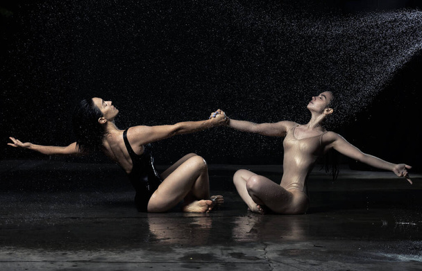 Дві вродливі жінки кавказького вигляду з темним волоссям сидять на асфальті у краплинах води на чорному тлі, тримаючись за руки і дивлячись вгору. Люди в костюмах - Фото, зображення