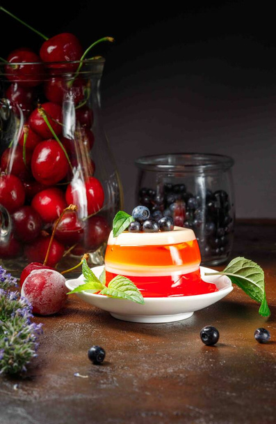 Natural berry jelly with a thin layer of milk, decorated with mint. A jug with cherries and blueberries in the background. Vertical, dark background, selective focus - Photo, Image