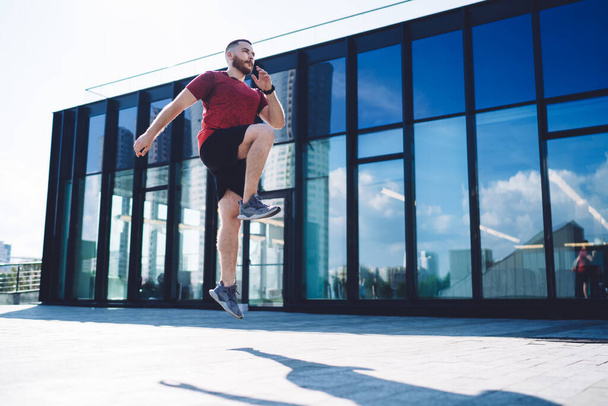 From below of full body young man in sneakers jumping on spot training and looking away while warming up during early summer morning - Фото, зображення