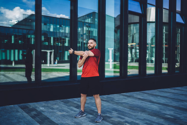 Full body of young bearded man with wristwatch training on pavement against modern building while looking away and enjoying early morning - Photo, image