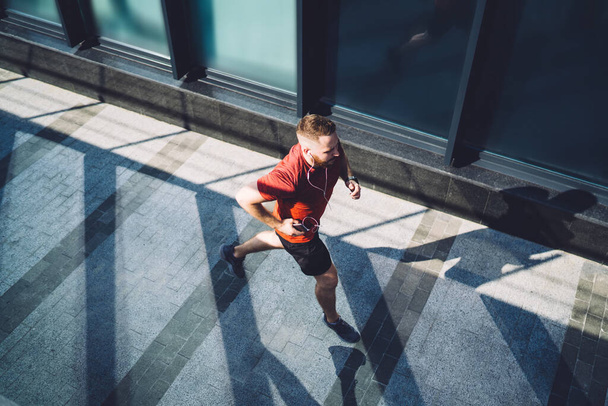 From above of full body athletic sportsman with phone running on pavement near glass wall and listening to music while looking away and enjoying morning - Foto, Imagen