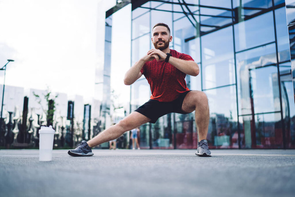 Ground level of muscular sportsman in red tee shirt looking away and doing stretching while warming up in city during early morning - Foto, Bild