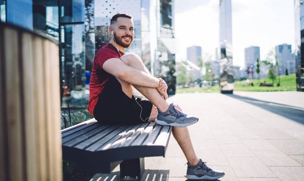 Side view of cheerful male athlete wearing sportswear listening to songs while sitting on bench and relaxing on sunny summer day - Foto, imagen