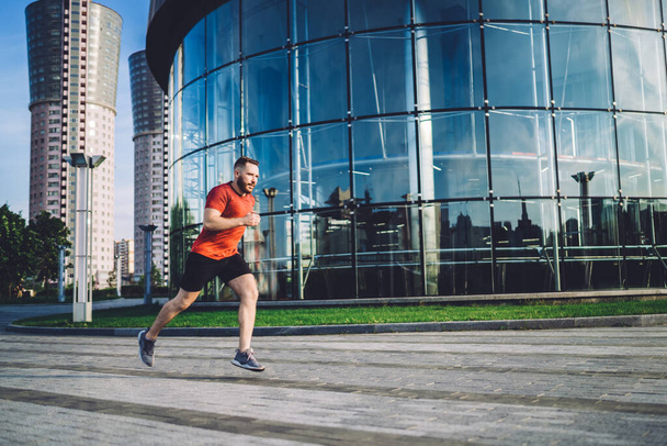 Full length of male athletic runner in sportswear running fast on pavement near modern glass building in city in summer sunny day - 写真・画像