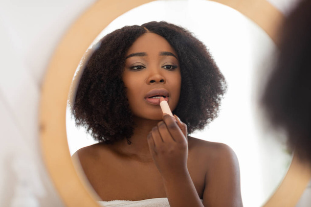 Ελκυστική νεαρή Αφρο κυρία εφαρμογή κραγιόν κοντά στον καθρέφτη στο σπίτι, βάζοντας διακοσμητικό μακιγιάζ. Ομορφιά και μόδα - Φωτογραφία, εικόνα