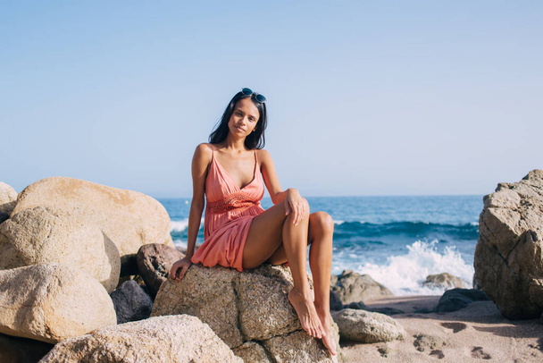 Portrait of beautiful Latin girl resting at coastline stone and looking at camera ready for sunbathing during tropical vacations, attractive female in sundress posing during pastime at seashore - Zdjęcie, obraz