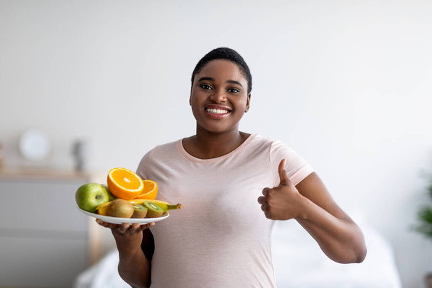 Feliz más tamaño mujer negra sosteniendo frutas y mostrando el pulgar hacia arriba, recomendando una dieta saludable para la pérdida de peso en casa - Foto, imagen