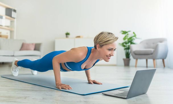 Femme gaie faisant planche sur tapis avec ordinateur portable à la maison - Photo, image