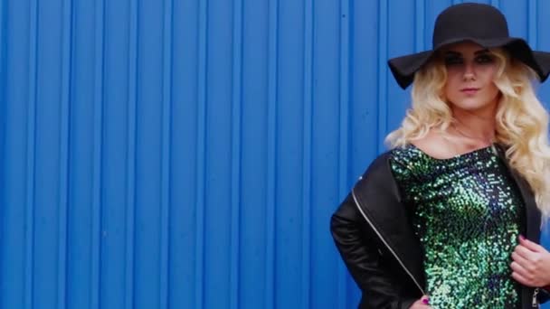 Krásná blondýnka v černém klobouku, černá kožená bunda, zelené flitrové šaty pózující na pozadí modré stěny. - Záběry, video