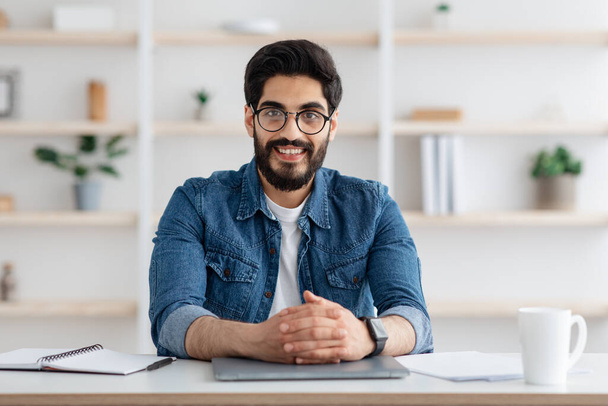 Szczęśliwy człowiek arabski samodzielny przedsiębiorca siedzi przy biurku w domu biura i uśmiecha się do kamery - Zdjęcie, obraz