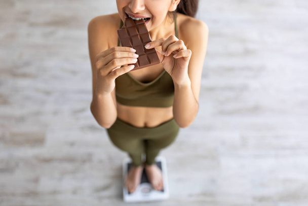 国会の失敗、食事の概念をカンニング。スケールに立っている間にチョコレートを食べるインドの女性のビューの上に - 写真・画像