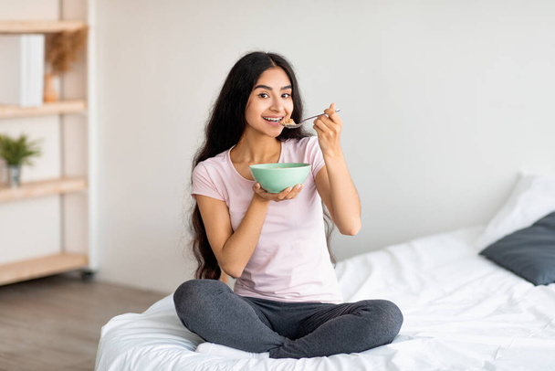 Belle dame indienne ayant des céréales avec du lait ou des flocons de maïs tout en étant assis sur un lit confortable à la maison - Photo, image