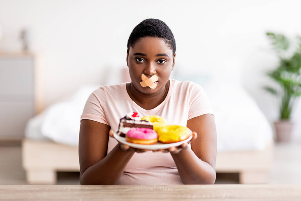 Nälkäinen plus koko musta nainen ottaa laihtuminen ruokavalio, yllään liima siteet suussa, tilalla levy makeisia - Valokuva, kuva