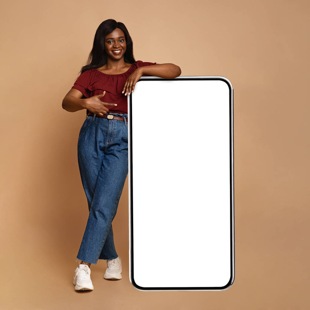Стильная черная женщина тысячелетия, указывающая на большой мобильный телефон, макет - Фото, изображение