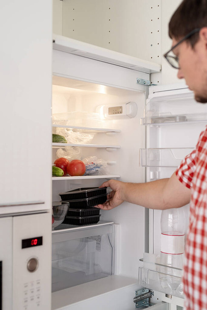 Delivery food, products to home. Shopping and healthy food concept. Young man in red plaid shirt holding disposable plastic boxed with food and putting them to the refridgerator - Photo, image