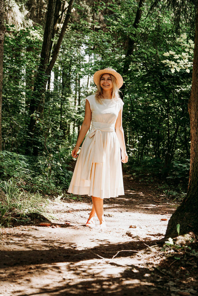 A girl in a white dress and hat walks through a summer park or forest - Fotó, kép