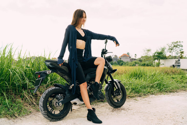 From below full body of happy young slender woman in cropped top and long summer cardigan sitting on bike and looking away with smile while having rest - Foto, afbeelding