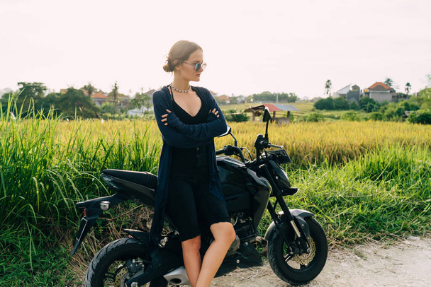 Serious young slender woman in stylish black clothing and sunglasses standing next to bike and looking away while having break - Zdjęcie, obraz